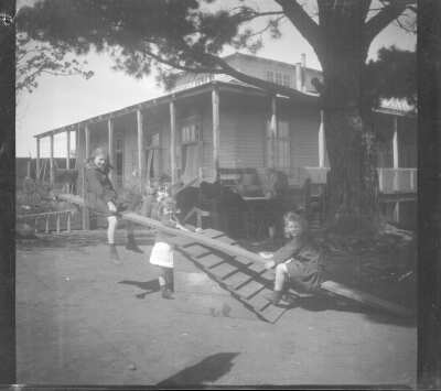 Plano general de una mujer adulta al fondo, junto a cinco niños y niñas jugando en el patio de una casa. Tres de las niñas juegan en un balancín.