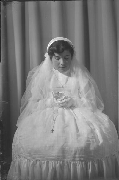 Retrato estudio plano entero de niña con Vestido de primera comunión sentada con una biblia en sus manos, posando de frente