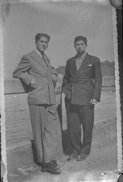 Reproducción de fotografía. Retrato de plano entero de dos hombres con Traje, uno de ellos con un cigarrillo en la boca, afirmados de una baranda en un lugar no identificado